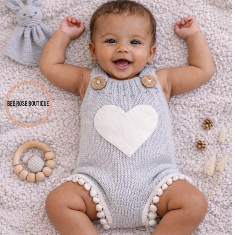 May include: A baby wearing a light blue knitted romper with a white heart applique and wooden button details. The romper has white pom-pom trim. A wooden teether and a stuffed bunny are nearby. The text "BEE ROSE BOUTIQUE" is in a circle.