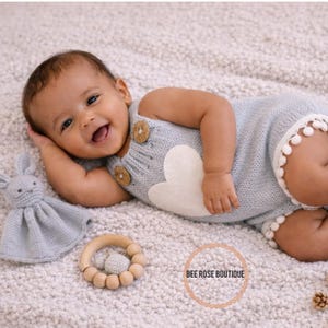 May include: A baby wearing a light blue knitted romper with a white heart design and wooden buttons. The romper has white pom-pom trim. A knitted bunny and a wooden teether are nearby. The baby is smiling and lying on a soft, textured surface.