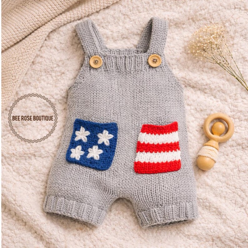 May include: A gray knitted baby romper with wooden button closures on the straps. The romper features two pockets, one with white stars on a blue background and the other with red and white stripes. A wooden rattle is next to the romper.