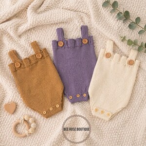 K&ouml;nnte beinhalten: Drei gestrickte Baby-Strampler in Senfgelb, Lavendel und Creme, jeweils mit Holzkn&ouml;pfen. Die Strampler haben einen gewellten Ausschnitt und Tr&auml;ger. Ein Holzherz und ein Bei&szlig;ring sind ebenfalls im Bild. Der Text "BEE ROSE BOUTIQUE" befindet sich in einem Kreis.