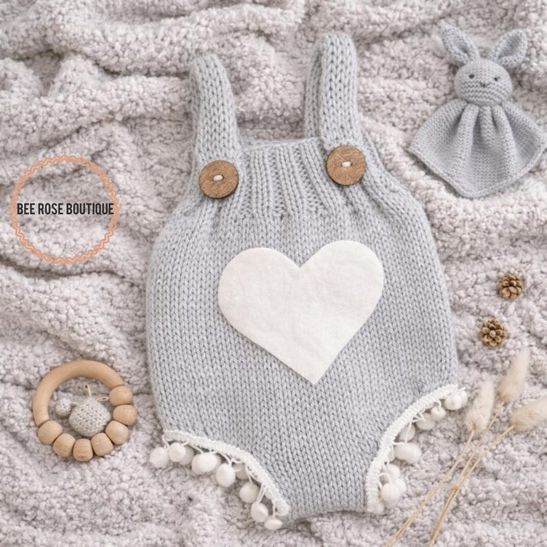 May include: A gray knitted baby romper with a white heart applique and wooden buttons. The romper has white pom-pom trim and is accompanied by a matching bunny lovey and a wooden teether. The text "BEE ROSE BOUTIQUE" is in the image.