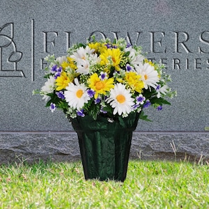 May include: A bouquet of yellow and white daisies with purple accents in a black plastic vase. The flowers are placed on a gray headstone with the text "Flowers & Funerals" on it.