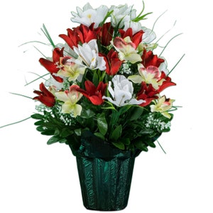 May include: A vibrant floral arrangement featuring red, white, and yellow artificial flowers. The bouquet includes lilies and other blooms, set in a dark green textured pot. Green leaves and sprigs add to the natural look.