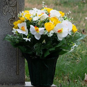 May include: A floral arrangement in a dark green pot, featuring white orchids, yellow roses, and small white flowers. The arrangement is placed near a gray headstone. The flowers are artificial and provide a colorful display.
