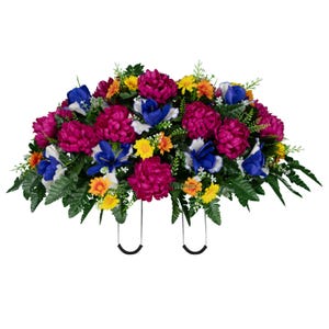 May include: A vibrant floral arrangement featuring large, magenta chrysanthemums, blue irises, and yellow and orange accent flowers. The arrangement is set against a backdrop of green foliage and includes two black metal support brackets.