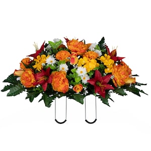 May include: A vibrant floral arrangement featuring orange peonies, red lilies, yellow daisies, and green foliage. The artificial flowers are arranged in a horizontal shape with two black metal supports, suitable for a graveside display.