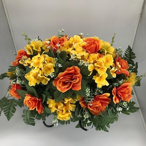 May include: A vibrant floral arrangement featuring orange roses and yellow hydrangeas, accented with green foliage and small white flowers. The arrangement is set in a black metal base, creating a colorful display.