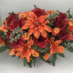 May include: A vibrant floral arrangement featuring a mix of orange dahlias, red roses, and yellow-orange tulips. Green foliage and ivy accents complement the colorful blooms. The arrangement is a mix of artificial flowers.