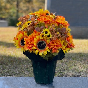 May include: A vibrant floral arrangement featuring sunflowers and orange blooms. The bouquet is presented in a dark green, textured container, creating a striking contrast with the bright colors of the flowers. The arrangement is set outdoors.