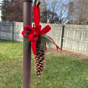 May include: A red ribbon bow is tied around a brown pine cone, hanging from a metal hook on a brown pole. The pine cone is decorated with green pine needles.