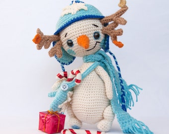 May include: A crocheted snowman figure with a blue hat, scarf, and a carrot nose. The snowman holds a small blue creature with candy canes and a red gift box. A candy cane lies on the ground. The snowman has brown antlers.