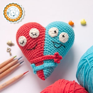 Puede incluir: Un juguete de peluche con forma de corazón tejido a crochet con una cara roja y azul. El juguete tiene ojos saltones y una boca sonriente. El juguete está sentado sobre una superficie blanca con ovillos de lana y lápices.