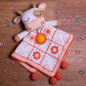 以下が含まれることがあります： オレンジとピーチのアクセントが付いた白い編み物のベビーブランケット。ブランケットには、目を閉じた編み物の牛の玩具と花のデザインが施されています。