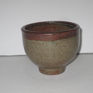 May include: A small, round ceramic bowl with a speckled, earthy green and brown body. The upper rim features a distinct, smooth band of dark reddish-brown glaze that contrasts with the textured, mottled finish of the lower portion of the bowl.
