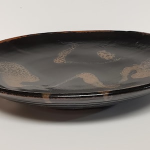 May include: A shallow, oval-shaped ceramic bowl with a glossy black glaze. The interior surface features abstract, textured brown and tan markings that appear as organic, mottled patches scattered across the dark, reflective base of the dish.