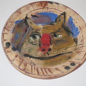 May include: A round ceramic plate featuring a hand-painted illustration of a cat face. The cat is rendered in shades of brown with a prominent red nose, green eyes, and black whiskers. The background includes blue accents, and the rim has brown markings.