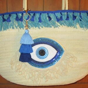 May include: A straw tote bag with a blue fringe trim and a large, embellished eye design. The bag features a blue tassel charm and woven handles. The eye design has a white center, blue sequins, and a dark blue border.