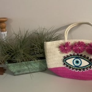 May include: A white and pink straw tote bag with a blue and white evil eye design and pink pom poms.