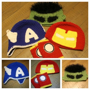 May include: Set of four crocheted hats featuring superhero characters. The hats are blue, green, red, and yellow with white accents. Each hat has a unique design, including a letter 'A', a green face with eyebrows, a red face with a yellow mask, and a yellow face with a red mask.