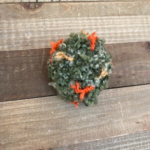 Pode incluir: Uma bola de fio feita à mão, texturizada, em tons de verde, laranja e bege. O fio é enrolado firmemente, criando uma forma esférica densa. Descansa sobre uma superfície de madeira.