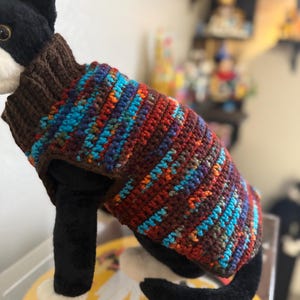 Puede incluir: Un suéter para gato multicolor tejido a mano. El suéter presenta un cuello alto marrón y un cuerpo con rayas rojas, azules, naranjas y verdes. El gato es blanco y negro, con una apariencia de felpa.