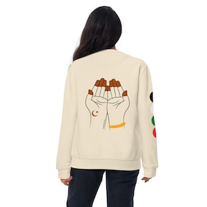 May include: A cream-colored sweatshirt with a graphic of two hands in a praying position on the back. The hands are brown with orange fingernails and a crescent moon and star on the left hand. The sweatshirt has a gold bracelet on the right wrist and a black, white, green, and red circle pattern on the right sleeve.