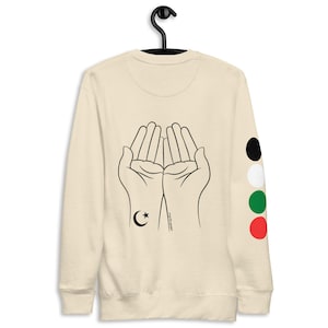 May include: A beige sweatshirt with a black outline of two hands in a praying position. The hands have a crescent moon and star on the wrist. The right sleeve has four colored circles: black, white, green, and red.