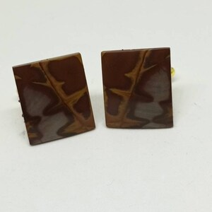 May include: A pair of square cufflinks with a brown, white, and orange marbled pattern. The cufflinks have a gold-colored metal backing.