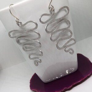 May include: A pair of silver earrings with a wavy, abstract design. The earrings are hanging from a white surface.