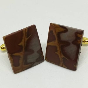 May include: A pair of square cufflinks with a brown, white, and gold pattern. The cufflinks are made of a smooth, polished material.