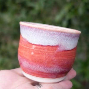 May include: A handmade ceramic cup with a rounded shape. The cup features a gradient of colors, with a white rim, transitioning to a reddish-orange body. The cup is held in a hand, showcasing its size and form.