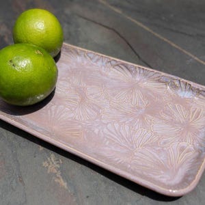 May include: A rectangular ceramic serving tray with a floral pattern in shades of pink and cream. Two green limes rest on the tray. The tray has rounded corners and a slightly raised edge.