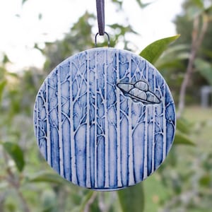 May include: Round ceramic ornament with a blue and white forest design and a small silver-colored UFO. The ornament is hanging from a dark gray ribbon. The background is blurred green foliage.