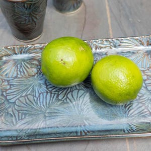 May include: Two bright green limes rest on a rectangular, blue-green ceramic tray with a floral pattern. The tray is next to two dark blue ceramic cups. The items are on a gray surface.