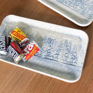 May include: A rectangular ceramic tray with a speckled white finish and a blue embossed forest pattern. The tray holds three small chocolate candy bars, including a Hershey's Milk Chocolate, a Mr. Goodbar, and a Krackel bar, resting on the surface.
