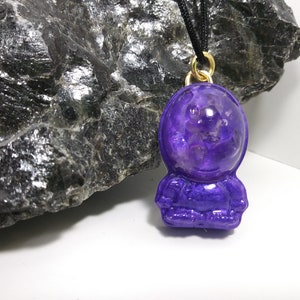May include: A purple astronaut charm necklace with a gold chain. The astronaut is wearing a clear helmet with a purple interior. The charm is hanging from a black cord.