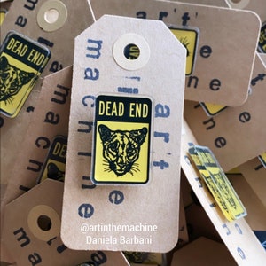 May include: A close-up of a stack of brown paper tags with the text "art" and "machine" printed on them. Each tag features a yellow and black enamel pin with the text "Dead End" and a graphic of a mountain lion's face.