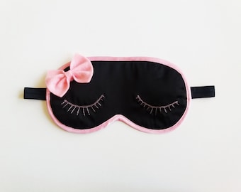 Può includere: Maschera per dormire nera con bordino rosa e un fiocco rosa. La maschera ha ciglia ricamate.