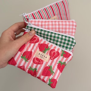 May include: A stack of four small, zippered pouches with various patterns. The top pouch has red and white stripes, the second has a pink gingham pattern, the third has a green gingham pattern, and the bottom pouch has a red strawberry pattern with white stripes. The pouches are labeled "EVESA designs."