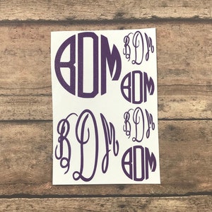 Pode incluir: Uma folha de papel branca com vários adesivos de monograma roxos em tamanhos diferentes. O monograma são as letras "BDM" em uma fonte de escrita.