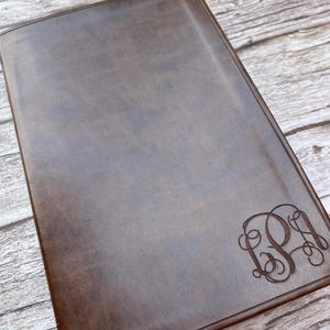 May include: A rectangular dark brown notebook with a smooth, matte finish. The front cover features a decorative, engraved monogram in the bottom right corner, consisting of three intertwined, cursive-style letters in a dark, recessed script.