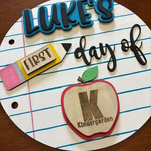 Op de afbeelding: Een rond houten bord met een schoolthema. Het bord heeft de tekst "LUKE'S day of" in zwarte en blauwe letters. Een potlood met het woord "FIRST" en een appel met de letter "K" en het woord "Kindergarten" zijn ook afgebeeld.