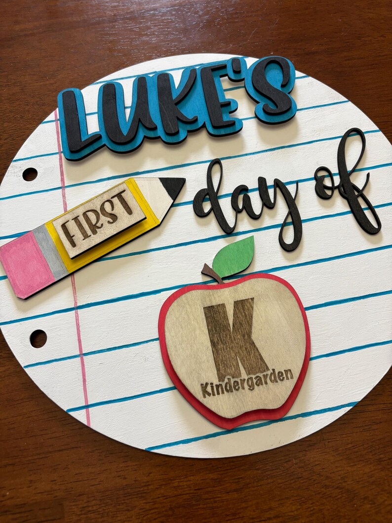 Op de afbeelding: Een rond houten bord met een schoolthema. Het bord heeft de tekst "LUKE'S day of" in zwarte en blauwe letters. Een potlood met het woord "FIRST" en een appel met de letter "K" en het woord "Kindergarten" zijn ook afgebeeld.