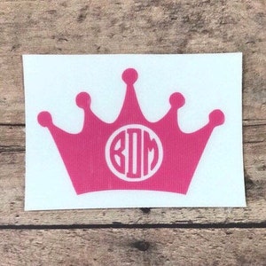 May include: Pink vinyl sticker of a crown with a white circle monogram in the center. The monogram reads "KDM".
