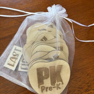 Op de afbeelding: Een verzameling hartvormige houten labels met de tekst "PK Pre-K" in bruin gedrukt. De labels zitten in een doorzichtige witte trekkoordzak. Een apart label leest "LAST".