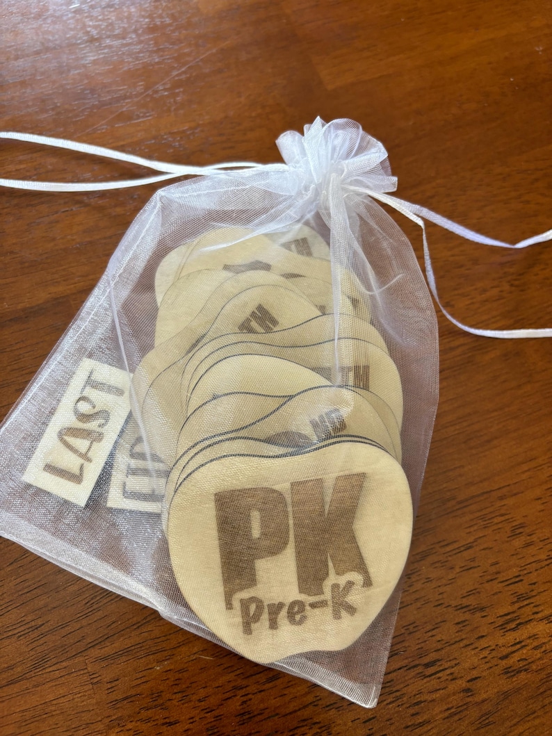 Op de afbeelding: Een verzameling hartvormige houten labels met de tekst "PK Pre-K" in bruin gedrukt. De labels zitten in een doorzichtige witte trekkoordzak. Een apart label leest "LAST".