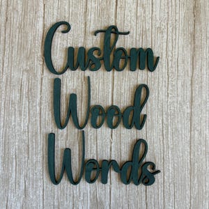 May include: Dark green wooden cutout letters arranged in three rows spelling the phrase Custom Wood Words. The letters feature a cursive script font with a smooth finish, showcasing the natural grain of the wood visible through the dark green paint.