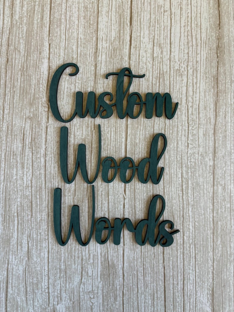 May include: Dark green wooden cutout letters arranged in three rows spelling the phrase Custom Wood Words. The letters feature a cursive script font with a smooth finish, showcasing the natural grain of the wood visible through the dark green paint.