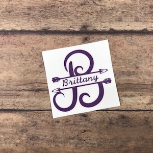 May include: Purple vinyl decal with the letter "B" and the name "Brittany" written across the middle of the letter. The letter is decorated with arrows on either side.