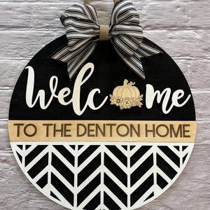 Op de afbeelding: Een ronde houten welkomstbord met een zwarte achtergrond en witte tekst "Welcome". Een decoratieve pompoen staat in het midden. De onderste helft heeft een zwart-wit chevronpatroon. Een jute lus en gestreepte strik staan bovenaan. De tekst "TO THE DENTON HOME" is ook inbegrepen.